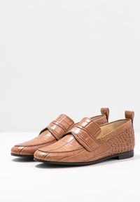 Brown loafers made of textured leather, featuring a smooth front and a patterned back. Velcro strap detail and low profile design.