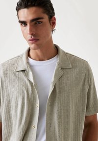 Short-sleeve, light beige shirt with textured diamond pattern, soft fabric, open collar, and two front button closures, layered over a white tee.