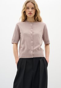 Kurzärmlige, rosafarbene Strickjacke mit Rundhalsausschnitt, sechs Knöpfen vorne und geripptem Saum, getragen über plissierten schwarzen Hosen.