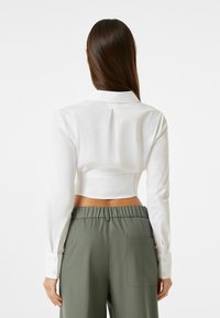 Camicia bianca cropped con maniche lunghe, caratterizzata da un dettaglio a pieghe sul retro. Abbinata a pantaloni verdi a vita alta con tasche laterali.