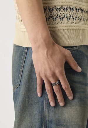 Sølv lenke-ring på en hånd som hviler på denim jeans. Hånden har en natural hudtone. Bakgrunnen er lys og nøytral.