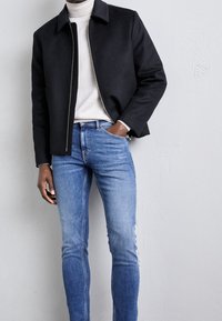 Černá zipová bunda s úzkým límcem na světle šedém roláku, v kombinaci s úzkými modrými džíny. Hladká tkanina s jemnou strukturou.