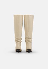 Cizme bej până la genunchi, din piele netedă, cu un design lejer și vârful ascuțit, având toc stiletto negru. Detalii de cusătură vizibile.