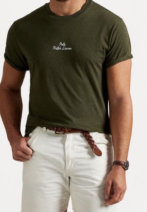 Homme portant un t-shirt Polo Ralph Lauren vert foncé à manches retroussées, un pantalon blanc, une ceinture tressée marron et une montre en cuir marron.