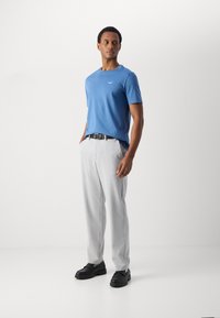Blauwe t-shirt met korte mouwen, lichtgrijze op maat gemaakte broek, zwarte riem en zwarte robuuste schoenen, staand op een witte achtergrond.