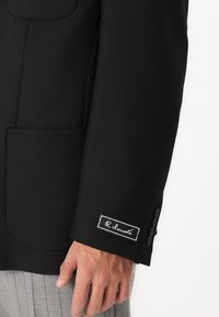 Μαύρο προσαρμοσμένο σακάκι με υφή, που διαθέτει τσέπη με καπάκι και ετικέτα επωνυμίας στο μανίκι. Ορατό διακοσμητικό κουμπί στον καρπό.