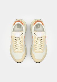 Beige og kremfargede atletiske joggesko med nettmateriale, oransje detaljer, teksturerte overflater og en svart gummisåle.