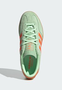Zielone zamszowe sneakersy z pomarańczowymi trzema paskami, teksturowanym wykończeniem i jaskrawym pomarańczowym logo na języku. Gumowa podeszwa ze wzorem w dwóch kolorach.