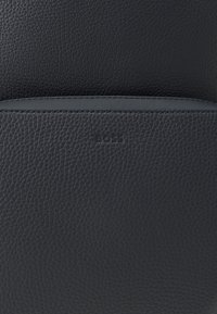 Artimas juodo tekstūruoto odos paviršiaus vaizdas, kuriame matosi siūlės ir subtiliai įspaustas "BOSS" logotipas.