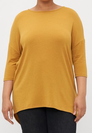 Kobieta w luźnej bluzce w kolorze musztardowym z rękawami do połowy przedramienia i czarnych spodniach, stojąca na tle jasnego, jednolitego tła.