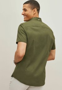 Camisa de manga corta en verde oliva, con cuello tipo botón y tejido suave. Corte entallado con un discreto detalle de costura en la parte posterior.