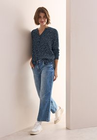 Marinblus med vitt abstrakt mönster, V-hals, långa ärmar; ljusblå raka jeans; vita sneakers, stående mot en vägg.