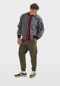 Siva bomber jakna s rebrastim rukavima, crvena majica, maslinaste cargo hlače i crne tenisice s bijelim detaljima. Ima aplikaciju i zipere.