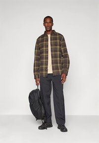 Karirana lanu s flanele v zelenih in rjavih odtenkih, bež majica, črne hlače in črni čevlji. Drži črn nahrbtnik.