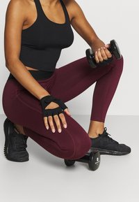 Atletka klečí v mahagonových legínách, černém tričku bez rukávů a rukavicích, drží jednoruční činku s další činkou na podlaze.