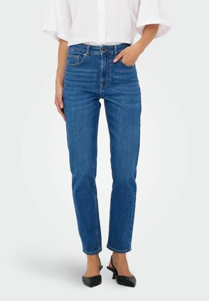 Kvinde iført blå jeans med lige ben, hvid løs skjorte og sorte slingback-sko med lav hæl, stående mod en ensfarvet baggrund.