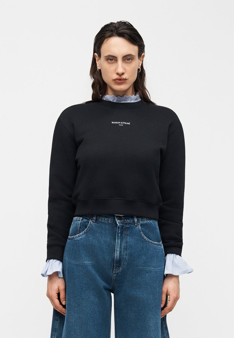 Γυναίκα που φοράει μαύρο φούτερ Maison Kitsuné πάνω από ανοιχτόγαλανη μπλούζα με βολάν, συνδυασμένο με ψηλόμεσο μπλε τζιν, στέκεται μπροστά σε λευκό φόντο.