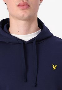 Homme portant un sweat à capuche bleu marine avec cordons et un petit logo d'oiseau jaune sur la poitrine, chemise blanche visible en dessous.