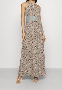 Robe maxi en tissu vert clair avec un motif floral rouge et blanc. Le corsage est smocké, avec un col halter.