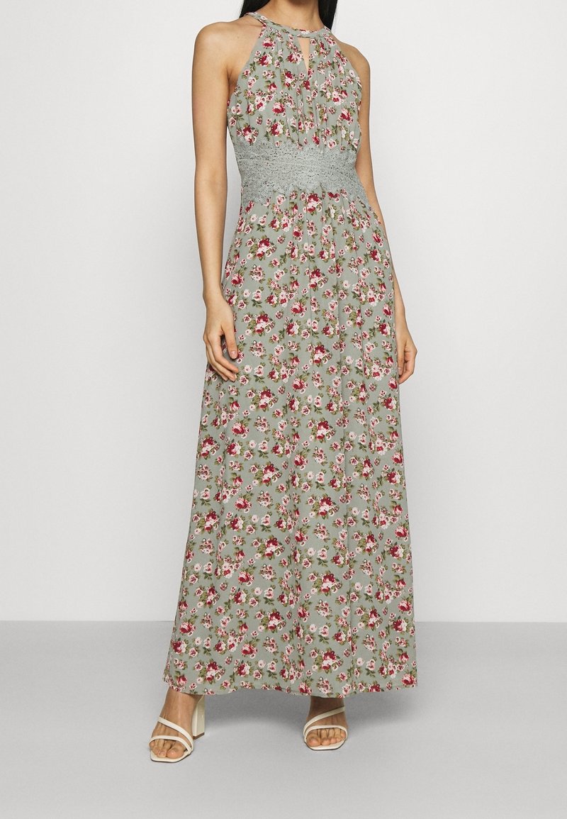 Robe maxi en tissu vert clair avec un motif floral rouge et blanc. Le corsage est smocké, avec un col halter.