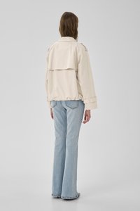 Person mit langen braunen Haaren, trägt eine beige Jacke und hellblaue ausgestellte Jeans, steht mit dem Rücken zur Kamera vor einem einfarbigen Hintergrund.