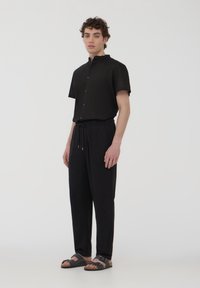 Camicia nera a maniche corte con colletto alla coreana, abbinata a pantaloni neri con coulisse e sandali grigi con cinghie regolabili.