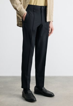 Mężczyzna w czarnych, dopasowanych spodniach, czarnych mokasynach, beżowej kurtce w kratę i żółtej koszuli, stojący na szarej podłodze na tle białej ściany.