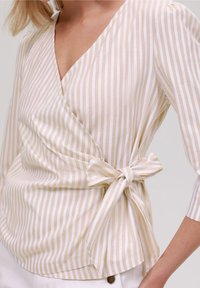 Páskový top v bílé a světle béžové barvě, se širokým výstřihem do V a bočním zavázáním. Vyrobeno z texturované, lehké látky.