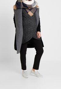 Grauer Umhang, dunkler Pullover, schwarze Hose und hellgraue Sneakers. Großes kariertes Tuch in Marineblau, Beige und Grau mit fransigen Kanten.