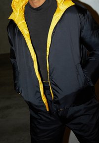 Svart pufferjacka med gul foder, med en framzipper och två sidofickor. Tyget ser slätt och lättviktigt ut.