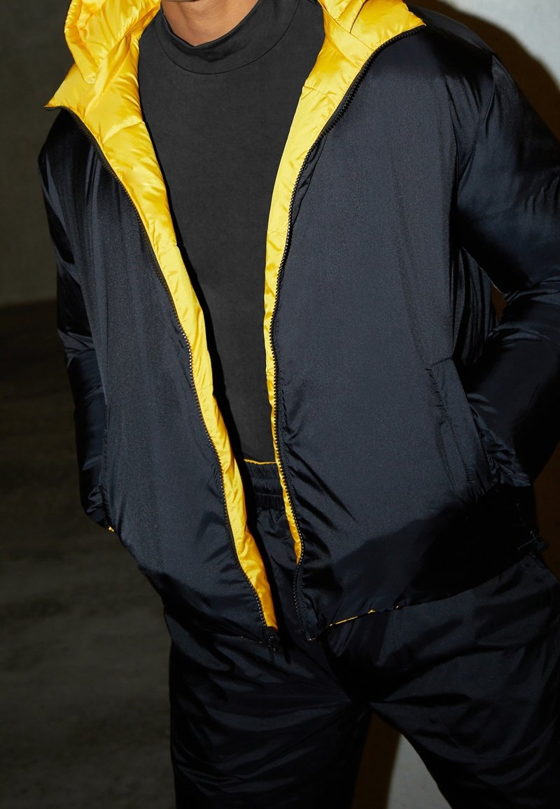 Svart pufferjacka med gul foder, med en framzipper och två sidofickor. Tyget ser slätt och lättviktigt ut.