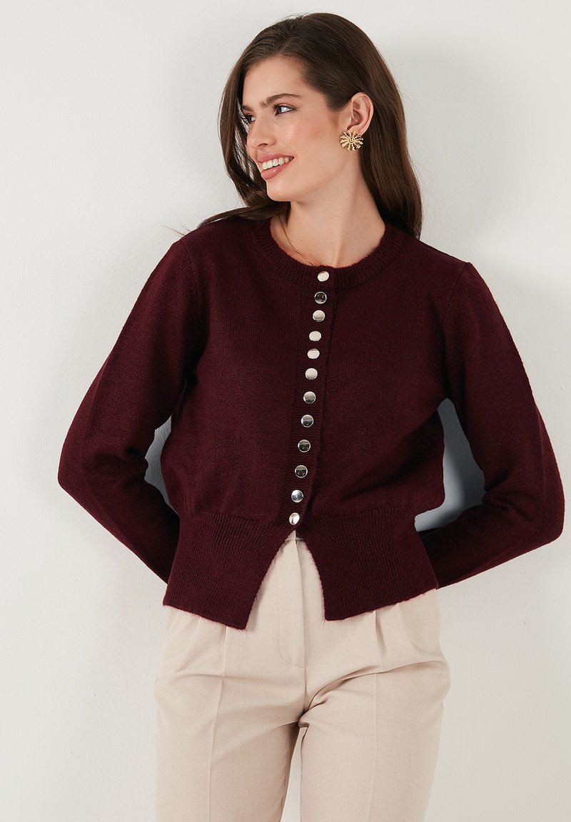 Cardigan en tricot bordeaux avec un col rond, huit boutons en argent sur le devant, et un design court avec un ourlet fendu à l'arrière.