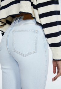 Jasnoniebieskie, dopasowane spodnie dżinsowe z wysokim stanem, wyposażone w klasyczny design z pięcioma kieszeniami i kontrastowym przeszyciem. Nad nimi widoczny pasiasty sweter z dzianiny.