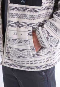 Giacca in pile bianca con motivi geometrici grigi, dotata di una tasca sul petto nera e tasche laterali. Tessuto morbido con chiusura a zip.