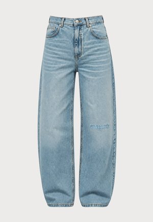 Femme portant un bonnet orange, un t-shirt rose oversize sur une chemise rouge rayée, un jean bleu ample et des baskets roses, debout devant un fond uni.