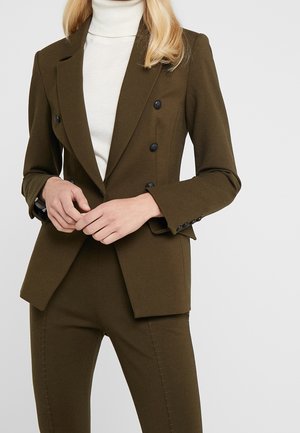 Vrouw met een olijfgroene double-breasted blazer en bijpassende broek, gecombineerd met een witte coltrui, handen voor zich geklemd.