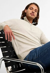 Beżowy sweter na zamek z miękkiego materiału, z prążkowanym kołnierzem i mankietami, w połączeniu z niebieskimi dżinsami, na czarnej plecionej krześle.