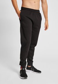 Schwarze Jogginghose mit einem elastischen Bund, seitlichen Taschen und gerippten Bündchen. Kombiniert mit schwarzen Sportschuhen mit weißen Akzenten.