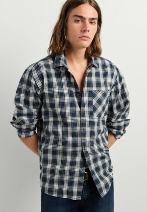 Junge Person mit langen braunen Haaren, die ein grün-blau kariertes Hemd mit hochgekrempelten Ärmeln vor einem schlichten hellen Hintergrund trägt.