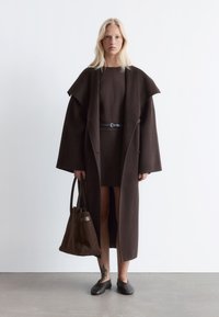 Brauner Wollmantel mit weit geschnittenen Ärmeln, über einem passenden Kleid getragen. Begleitet von einer braunen Tragetasche und Slippern. Glatte Textur.