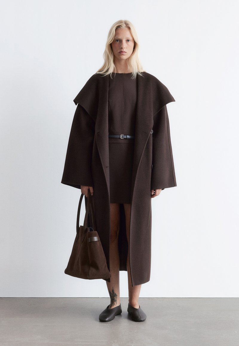 Brauner Wollmantel mit weit geschnittenen Ärmeln, über einem passenden Kleid getragen. Begleitet von einer braunen Tragetasche und Slippern. Glatte Textur.