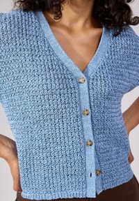 Ljusblå stickad cardigan med V-ringning, korta ärmar och stora knappar. Har ett texturerat, öppet stickmönster och lätt drapering.