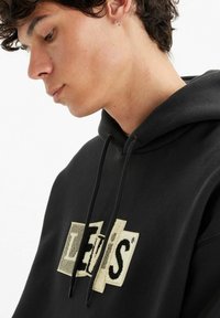 Black hoodie with a large textured "Levi's" logo in cream and black. Features a drawstring hood and soft material.