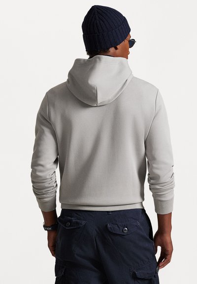 Hoodie cinza claro com gola cowl e punhos canelados, combinado com calças cargo azul escuro com bolsos com botão. As costas do modelo são visíveis.