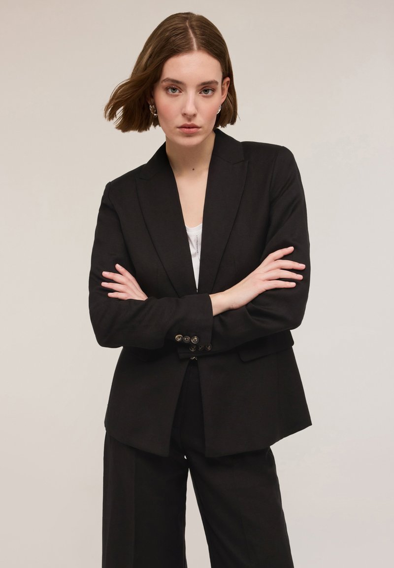 Czarna marynarka o dopasowanym kroju, z klapami i z guzikami przy rękawach; w połączeniu z białą bluzką i czarnymi spodniami. Gładka struktura.
