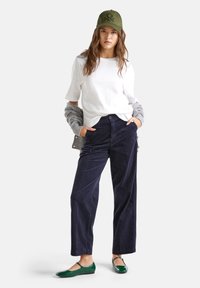 Young woman wearing olive green cap, white t-shirt, navy blue corduroy pants, gray sweater over arms, and green ballet flats, standing with hands in pockets.