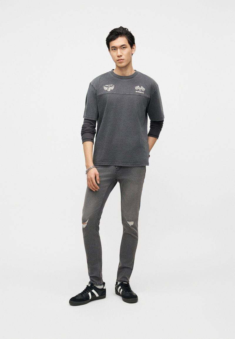 Young man standing wearing dark gray layered long sleeve and short sleeve t-shirt, ripped gray skinny jeans, and black sneakers.