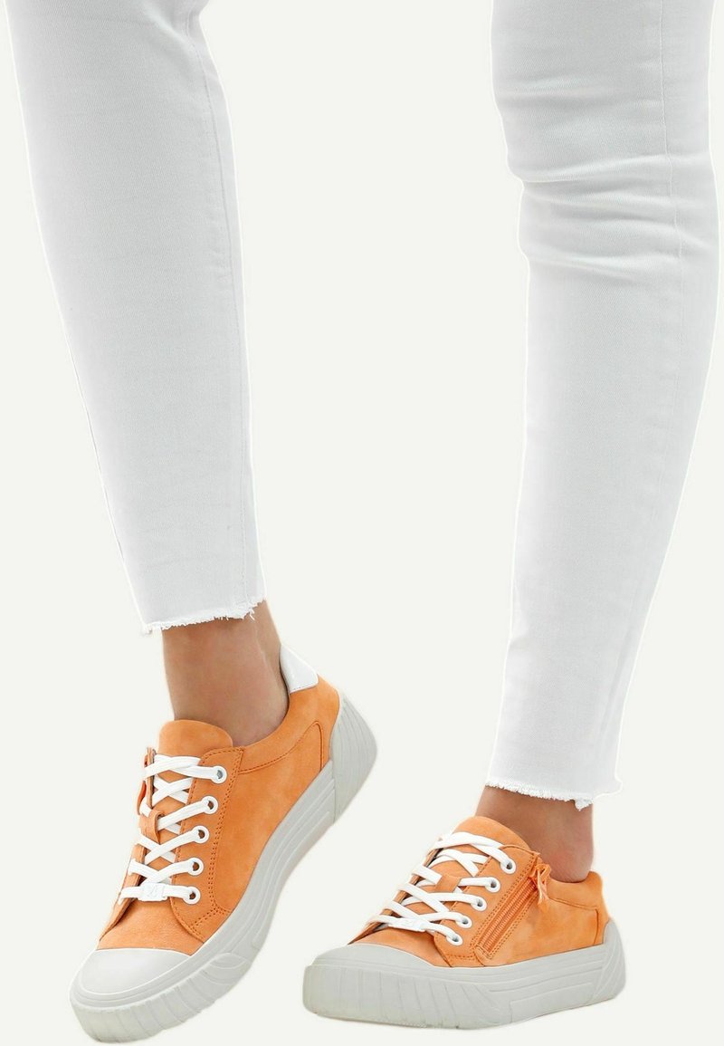 Person wearing white jeans and orange low-top sneakers with white laces and thick white soles, one foot slightly raised.