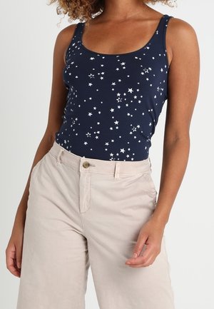 Navy Tanktop mit weißem Sternenmuster, aus weichem Stoff; kombiniert mit hellbeigen, hoch taillierten Hosen mit einem vorderen Knopf.