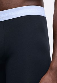 Črni bombažni bokserice z lahnim modrim elastičnim pasom, ki nosi logo "Calvin Klein". Gladka tekstura z oprijetim dizajnom.